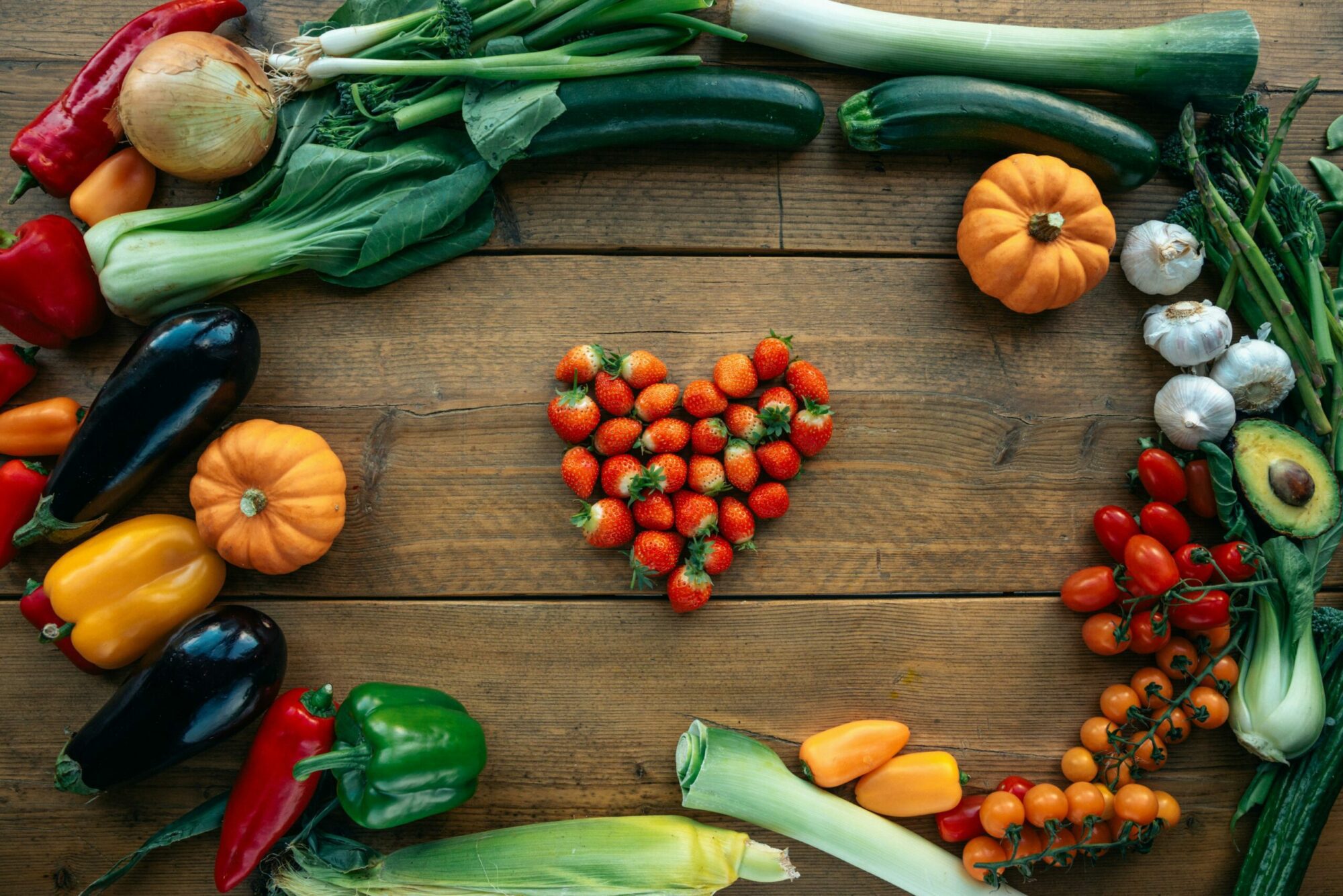 Un cœur en forme de fraise est placé au centre d'une table en bois, entouré d'un assortiment de légumes colorés, dont des poivrons, des aubergines, des tomates, des mini-citrouilles, des poireaux, de l'ail, du maïs, du bok choy, des oignons et des courgettes, disposés dans un cadre ovale lâche.